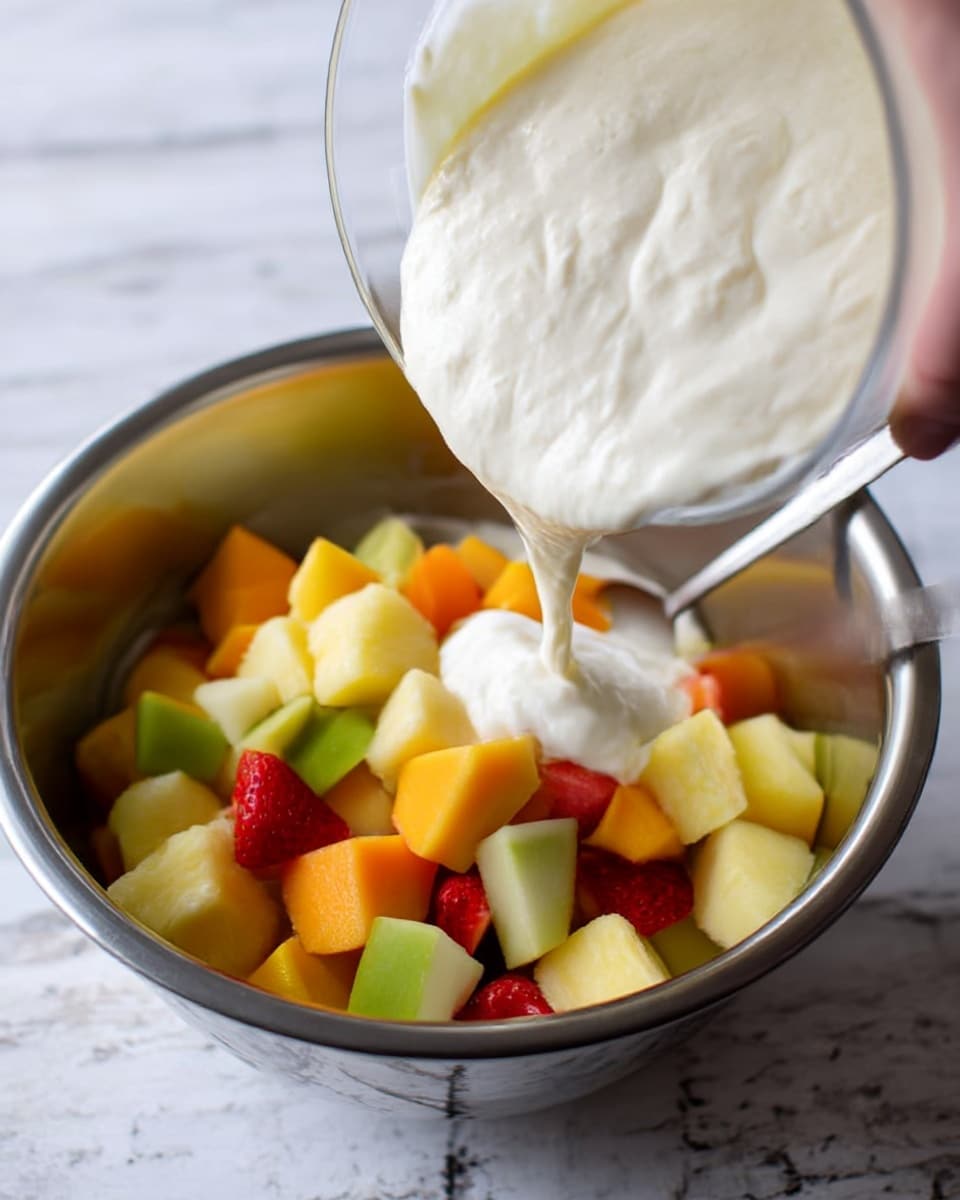 The image shows a close-up view of a clear bowl held by woman's hand pouring a thick, creamy white mixture over a bowl filled with colorful chopped fruit including cubes of yellow mango, green apple, orange pieces, pineapple, and red strawberries. The fruit bowl is metallic and has a shiny silver look, with a spoon resting inside it. The background texture is changed to a white marbled surface. photo taken with an iphone --ar 4:5 --v 7