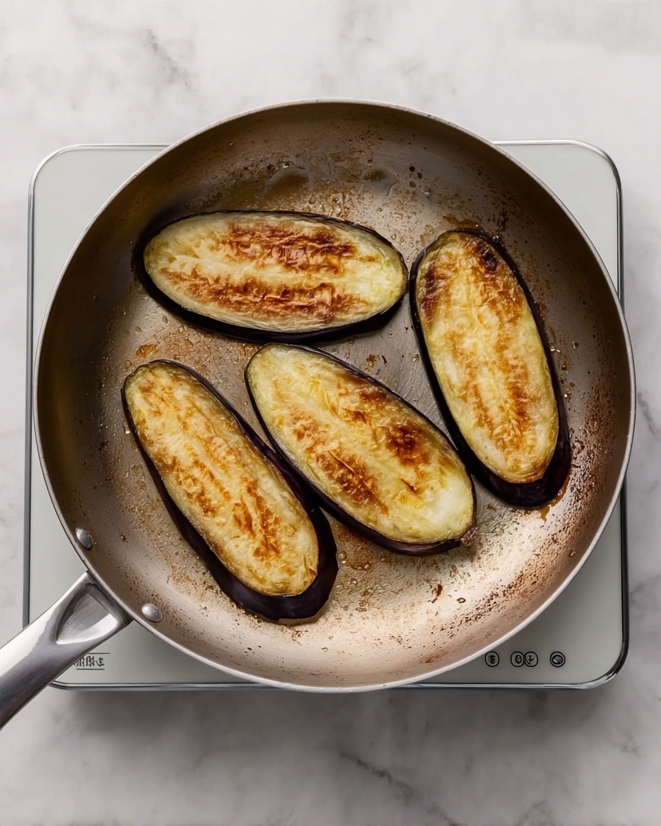 The image shows a frying pan on a white marbled surface with four slices of eggplant inside. Each eggplant slice is long and oval-shaped, with a dark purple skin around the edges and a golden-brown, slightly charred surface in the middle. The pan has a metallic look with some browning marks inside, and it sits on a white induction cooktop with simple buttons visible at the bottom. The overall colors include deep purple, light brown, and off-white with the pan and cooktop's metallic tones. photo taken with an iphone --ar 4:5 --v 7