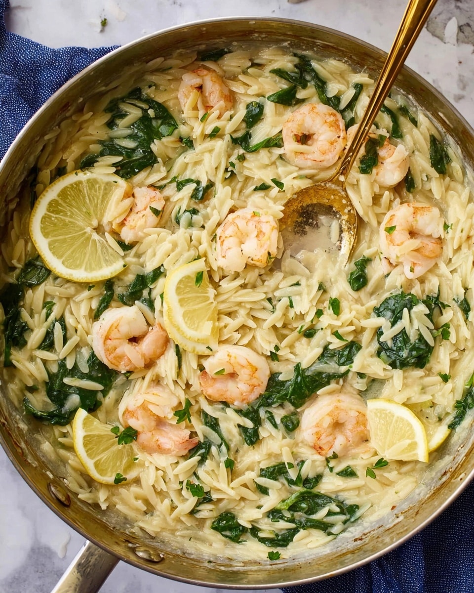 A round metal pan filled with creamy orzo pasta mixed with green spinach leaves spread evenly throughout the dish. Scattered on top are six pinkish-white shrimp, some lightly curled and partially covered with the sauce. Two thin lemon slices rest on the surface, one near the center and one near the edge. A gold spoon is placed inside, partially submerged in the orzo, and a small splash of sauce clings to its handle. The background is a white marbled surface with a blue cloth partially visible under the pan. Photo taken with an iphone --ar 4:5 --v 7