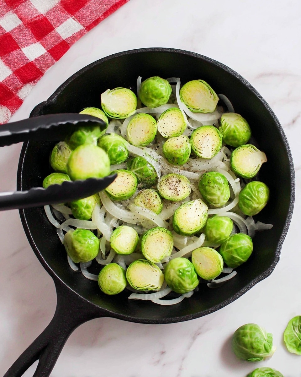 A black cast iron pan holds a single layer of bright green halved Brussels sprouts, facing cut side up, scattered over a bed of thinly sliced translucent onions with a light sprinkle of black pepper on top, all positioned near the center of the pan. The pan sits on a white marbled surface with a red and white checkered cloth partially visible in the upper left corner. A pair of black tongs holds one Brussels sprout toward the left edge of the pan, and a few loose Brussels sprout leaves rest on the white marbled surface near the bottom right corner. Photo taken with an iphone --ar 4:5 --v 7