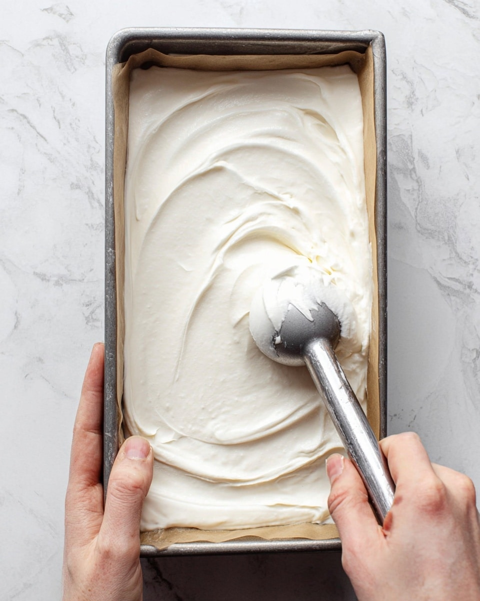 A rectangular metal tray lined with light brown parchment paper is filled with a smooth, thick white cream layer being spread evenly by a metal ice cream scooper held by a person's fingers. The cream layer is thick and soft, with gentle swirls and slight indentations from the scooper. The tray is held by a person's other fingers from the side against a background of white marbled texture. photo taken with an iphone --ar 4:5 --v 7