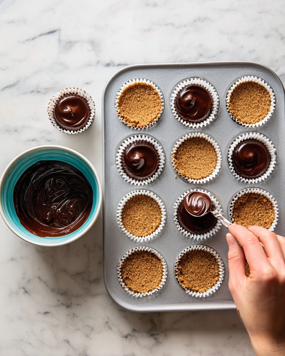 A silver muffin tray with white paper liners holds a layer of light brown crumbly crust in each cup. On top of the crust, a dark rich chocolate filling is being spooned in by a woman's hand from a small shiny spoon. Nearby, a white bowl with a bright turquoise inside holds extra chocolate mixture. The whole scene is set on a white marbled surface. photo taken with an iphone --ar 4:5 --v 7