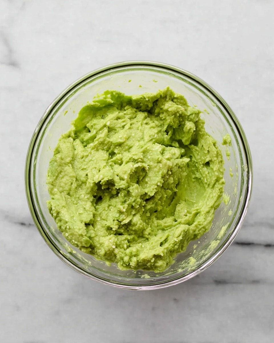 A clear glass bowl filled with a single layer of chunky, light green mashed avocado spread evenly inside. The texture shows small lumps throughout, giving it a thick, creamy look with some unevenness on the surface. The bowl is placed on a white marbled surface, making the green color of the avocado stand out brightly. photo taken with an iphone --ar 4:5 --v 7