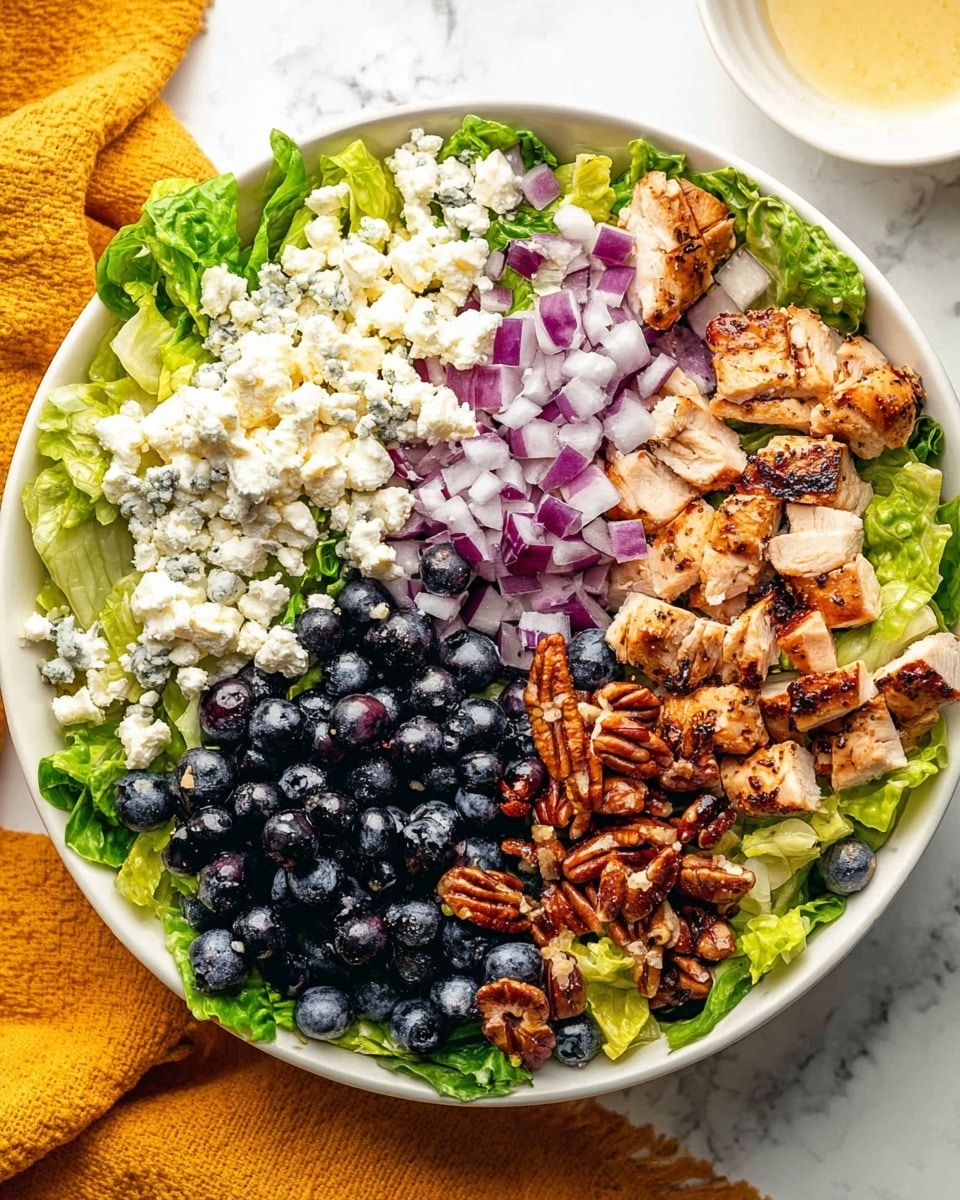 A white bowl filled with a colorful salad laid out in separate sections: at the base, green leafy lettuce with a crisp texture covers the bottom. On the top right, there are grilled, lightly browned pieces of chicken with a slightly charred look. To the left of the chicken, there are small white crumbles of cheese scattered over the salad. Below the cheese, there are pieces of chopped pecans with a glossy brown shell texture. Next to the pecans, a mix of fresh dark blue blueberries is piled up, showing a shiny surface and round shape. Above the blueberries, diced purple-red onions add a sharp contrast with their smooth texture. The bowl sits on a white marbled surface with a mustard-yellow cloth nearby and a cut white bowl with light dressing at the top right. Photo taken with an iphone --ar 4:5 --v 7