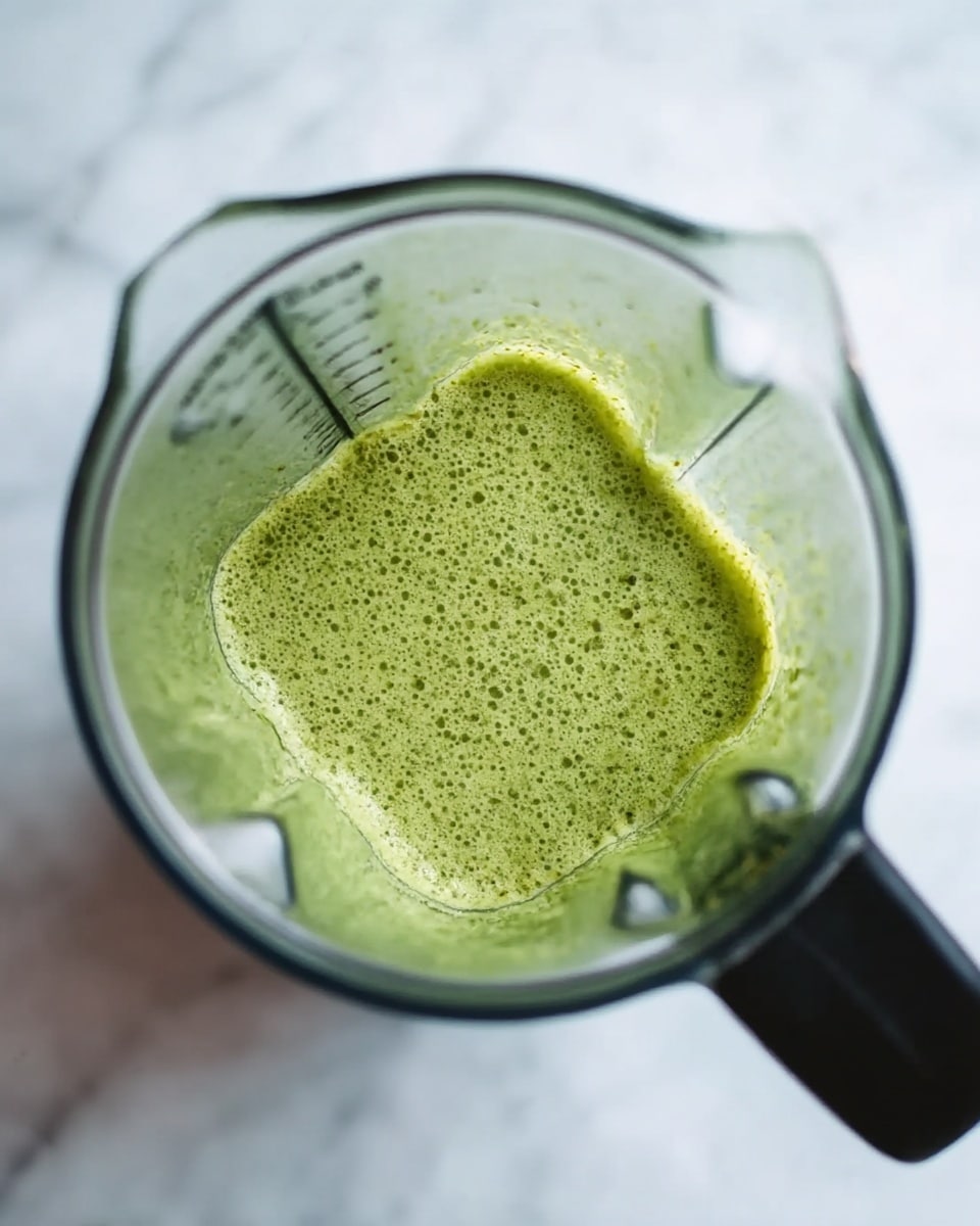 The image shows a close-up top view of a blender jar filled with a green liquid mixture that has a slightly frothy and textured surface. The blender jar is transparent with measurement markings on the side, and the handle is black. The background is a white marbled surface. Photo taken with an iphone --ar 4:5 --v 7
