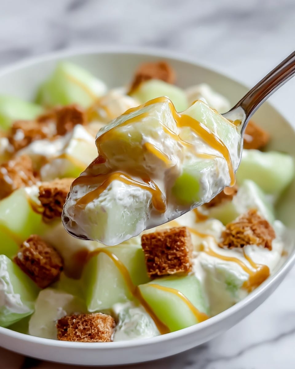 A close-up of a white bowl filled with a creamy salad showing three layers: large light green melon chunks with smooth, shiny texture; small brown bread cube pieces with a rough, crumbly texture; and white creamy dressing holding everything together with a smooth, thick texture, drizzled with thin lines of golden caramel sauce on top. A spoon lifted towards the camera holds a mix of these three layers, highlighting their colors and textures against a white marbled surface. photo taken with an iphone --ar 4:5 --v 7