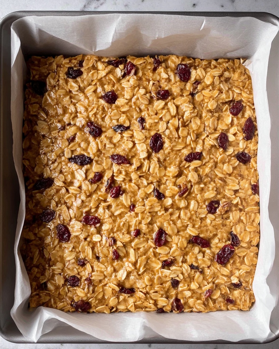 A single thick layer of golden oat mixture filled with visible dried red berries spreads evenly in a square baking tray lined with white parchment paper. The oats look slightly sticky and clumped together with a glossy texture, while the scattered dark red berries add spots of color throughout. The white marbled surface is partly visible around the edges of the tray. Photo taken with an iphone --ar 4:5 --v 7