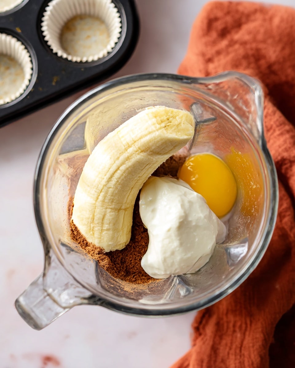 A clear glass blender container holds several ingredients that sit in separate sections at the bottom. There are three peeled bananas, pale yellow and slightly shiny, stacked on one side forming the largest layer. To the right, a dollop of thick white yogurt rests next to the bananas. Just above the bananas, a bright yellow raw egg yolk is visible. Underneath and around these, there is a brown powdery layer, likely cinnamon or cocoa, covering the base. The blender is placed on a white marbled surface, with a folded rust-colored cloth nearby and a black muffin tray with white paper liners mostly out of frame. Photo taken with an iphone --ar 4:5 --v 7