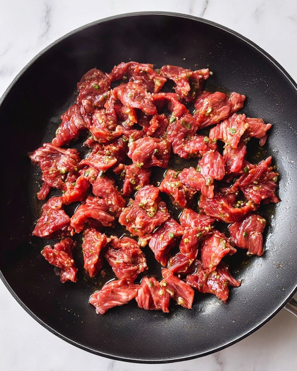 Thin slices of marinated raw beef with a reddish color mixed with small green herb bits are spread evenly across the bottom of a black frying pan. The beef pieces show some wetness and oil glistening, with slight steaming visible, and the pan surface has a smooth, shiny texture. The background is a white marbled surface. photo taken with an iphone --ar 4:5 --v 7