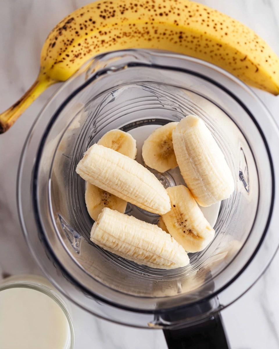 The image shows a close-up top view of a blender with five large pieces of peeled banana inside. The bananas are light yellow with soft textures and slight brown spots, resting on the metal blades at the bottom of the blender. Outside the blender, to the top left side, a whole ripe banana with many dark spots on its yellow peel is visible. The blender is placed on a white marbled surface, and in the bottom left corner, there is a partially visible glass of white milk. photo taken with an iphone --ar 4:5 --v 7