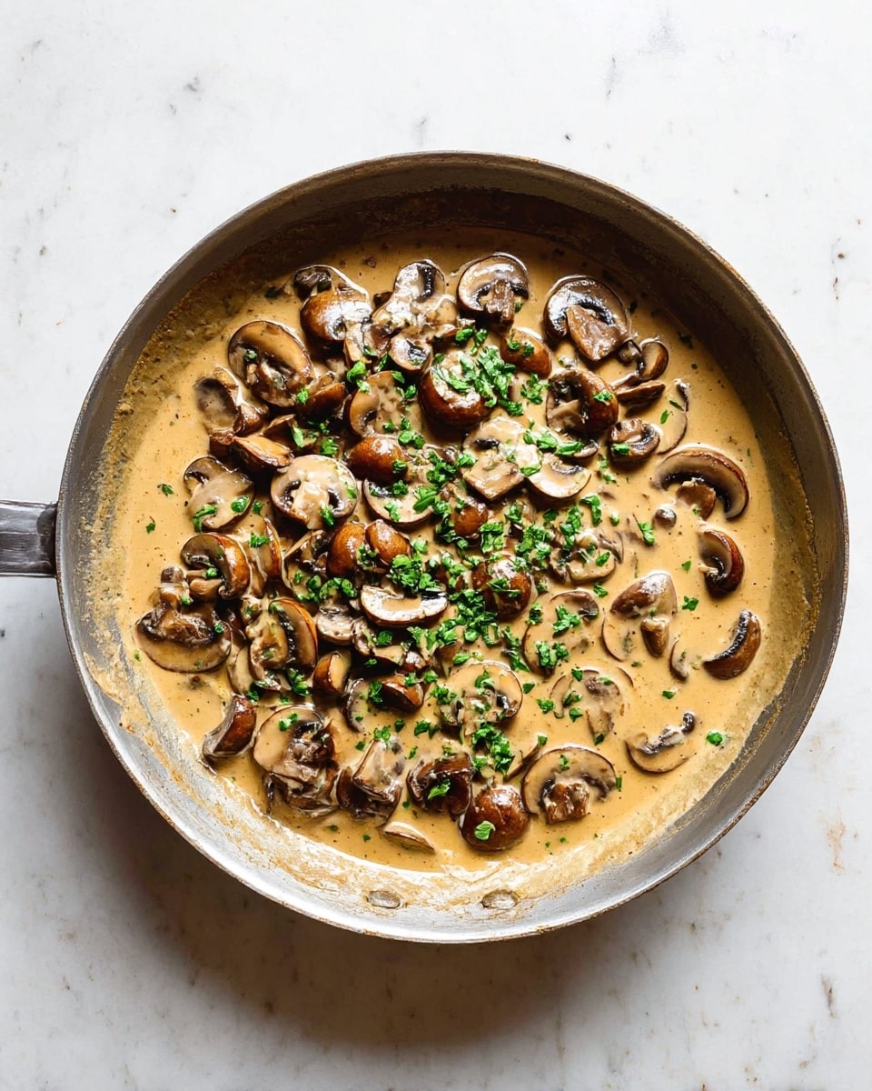 A gray metal pan filled with two main layers: the bottom layer is a creamy light brown sauce that spreads across the pan, while the top layer consists of many chopped brown mushrooms scattered inside the sauce. There are small green herb pieces sprinkled on top, adding a fresh touch. The pan rests on a white marbled surface. photo taken with an iphone --ar 4:5 --v 7