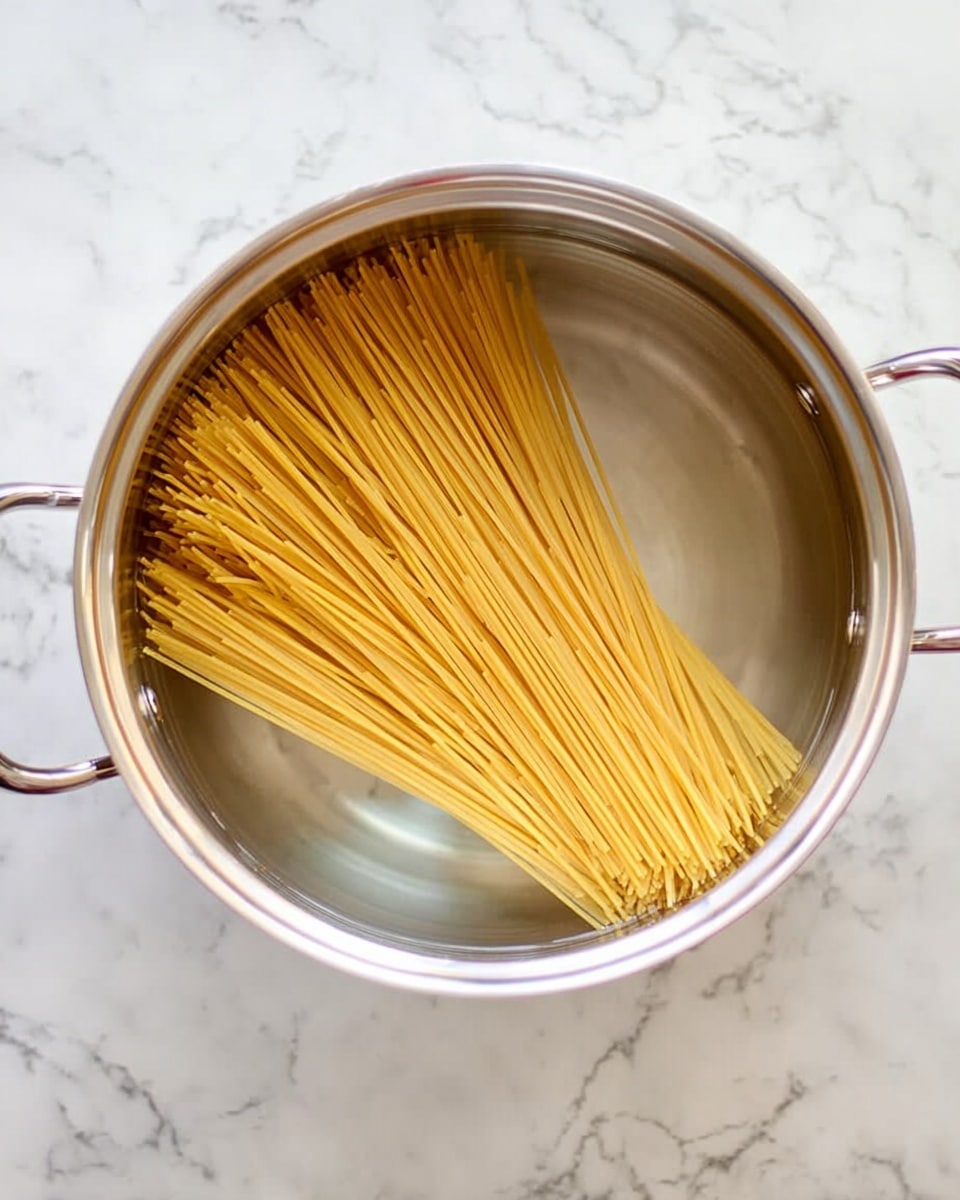 A shiny silver pot filled with clear water holds a large bunch of dry spaghetti placed diagonally inside. The spaghetti strands are yellow and do not yet bend in the water. The pot has two handles, one on each side. The pot sits on a white marbled surface. photo taken with an iphone --ar 4:5 --v 7
