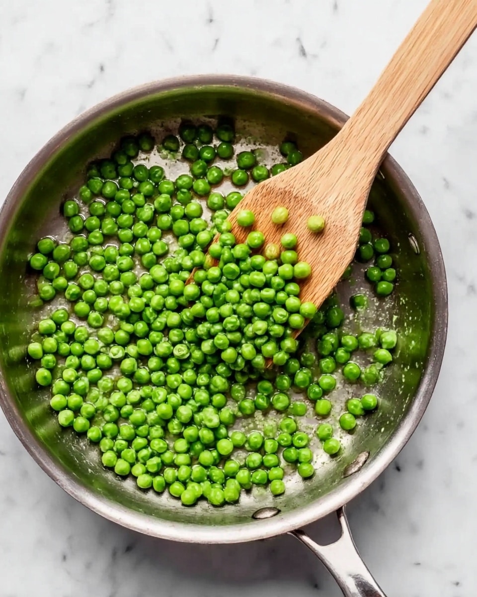 A close-up view of a steel pan with a shiny silver surface filled with bright green peas spread unevenly inside. A wooden spatula with a smooth light brown texture rests inside the pan on the right side, slightly lifting some of the peas. The pan sits on a white marbled surface, which adds a clean and fresh background to the scene. photo taken with an iphone --ar 4:5 --v 7