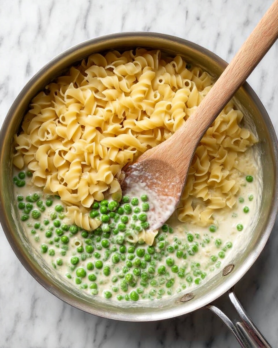 A silver pan on a white marbled surface holds a layered dish with three main visible parts: at the bottom, a creamy white sauce mixed with bright green peas; on top of that, a large pile of light yellow curled pasta tubes; a wooden spoon with a long handle rests in the pan, partially submerged in the sauce and peas. photo taken with an iphone --ar 4:5 --v 7