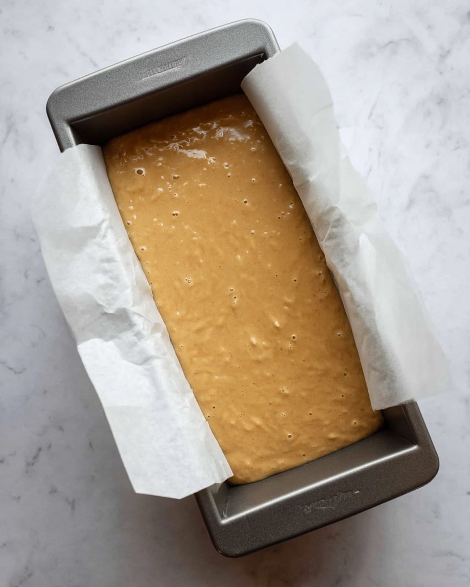A loaf pan lined with white parchment paper holds smooth, light brown batter that fills about three-quarters of the pan, with small bubbles and tiny lumps visible on the surface. The pan sits on a white marbled surface, and the batter has a soft, thick texture ready for baking. photo taken with an iphone --ar 4:5 --v 7