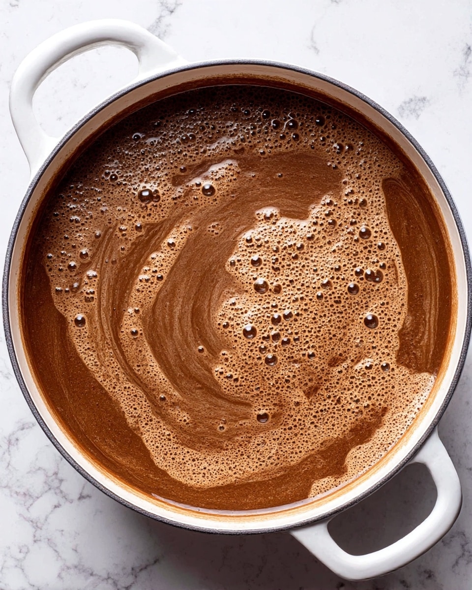 A large white cooking pot filled with a dark brown liquid that has a smooth surface with light swirls and small bubbles scattered across the top, creating a textured and frothy appearance. The pot has two white handles on opposite sides and sits on a white marbled surface. The liquid fills most of the pot, showing a rich, warm brown color with variations in tone from the swirls and bubbles. Photo taken with an iphone --ar 4:5 --v 7