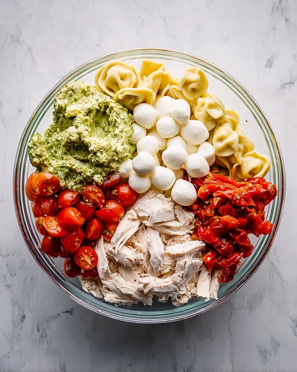 A clear glass bowl is placed on a white marbled surface, filled with five distinct layers of ingredients arranged in separate sections. One section holds creamy green pesto sauce with visible herbs and oil texture sitting on top of pale yellow tortellini pasta. Next to it, there are many small white mozzarella balls stacked on the pasta. Below this, bright red cherry tomatoes are halved, showing shiny, juicy interiors. Another section contains shredded white chicken meat with a soft, fibrous look. The final section has roasted red peppers displaying a glossy, slightly wrinkled surface. Photo taken with an iphone --ar 4:5 --v 7