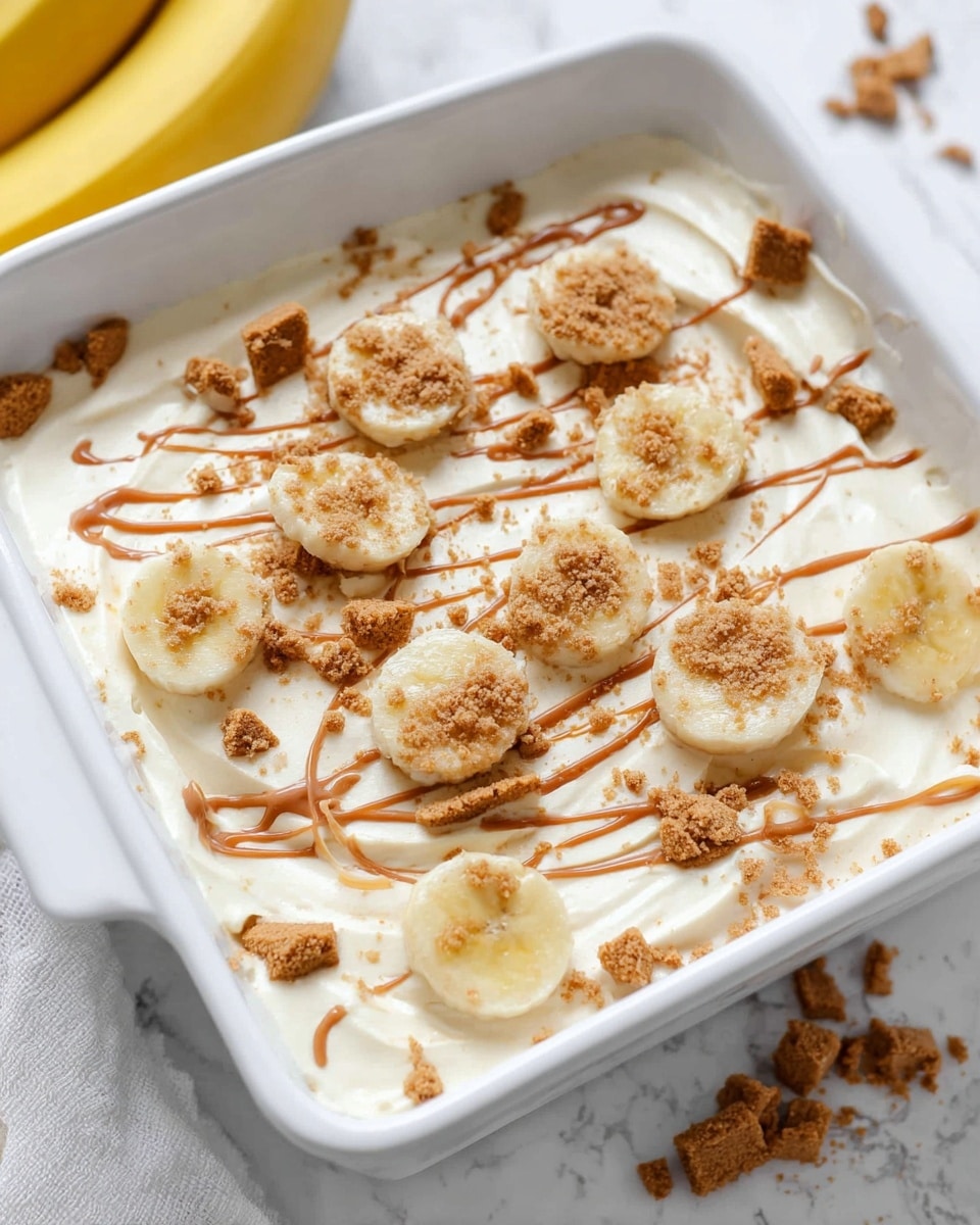 The image shows a white square dish filled with a creamy off-white layer that looks smooth and thick. On top, there are slices of pale yellow banana spread out unevenly, along with small clumps of light brown crumbled biscuits scattered across the surface. Drizzled over everything are light brown lines of sauce, adding a slightly shiny texture. The dish is set on a white marbled surface with a whole yellow banana partly visible on the left side. photo taken with an iphone --ar 4:5 --v 7