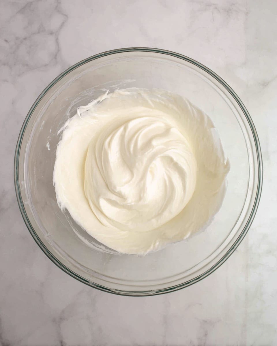 A clear glass bowl sits on a white marbled surface, filled with one smooth, thick layer of pale creamy white mixture that has a soft, swirled texture on top. The mixture looks light and fluffy with gentle curves and folds in the center. The bowl’s edges are thin and clear, showing the creamy content inside. Photo taken with an iphone --ar 4:5 --v 7