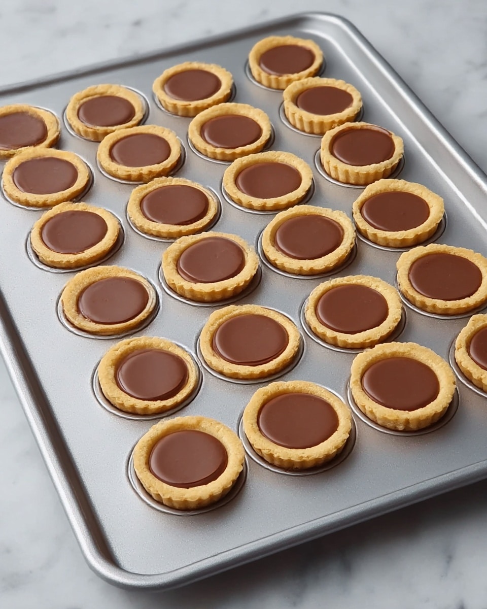 The image shows a metal baking tray with 24 small tartlets arranged in four rows and six columns. Each tartlet has two layers: a golden-brown tart shell forming the bottom and sides, and a smooth, round milk chocolate filling inside, slightly raised but even. The tray sits on a white marbled surface, with soft, even lighting highlighting the textures of the tart shells and chocolate filling. The tray's metal surface is slightly reflective, and the overall look is clean and neat. photo taken with an iphone --ar 4:5 --v 7