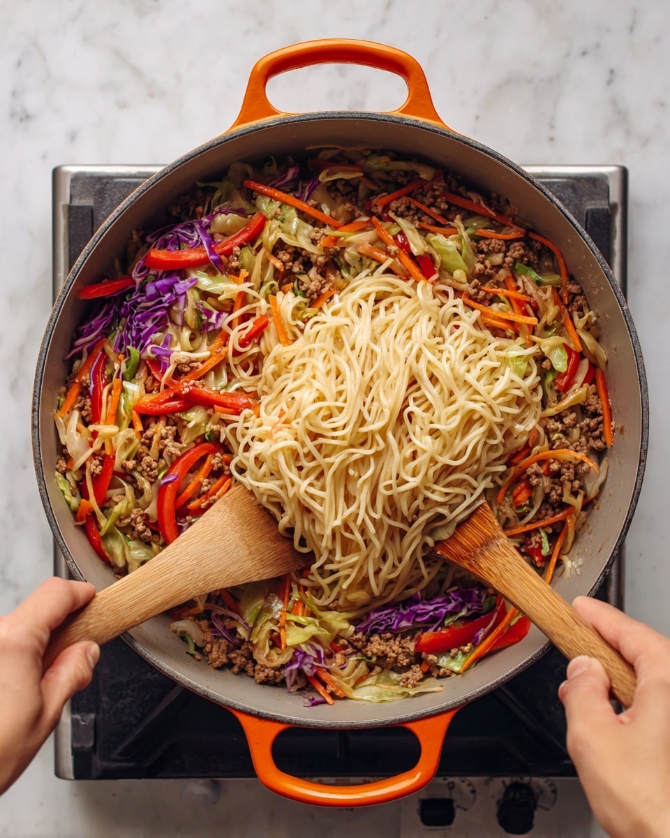 A large pot filled with cooked noodles sitting on top of colorful stir-fried vegetables and ground meat, including orange carrot strips, red bell pepper slices, and green cabbage pieces, all mixed with small bits of purple cabbage. Two wooden spatulas held by a woman's hands are lifting and mixing the noodles into the vibrant vegetable and meat base below. The pot has gray inside walls and orange handles, and it rests on a cooking stove set against a white marbled surface background. Photo taken with an iphone --ar 4:5 --v 7