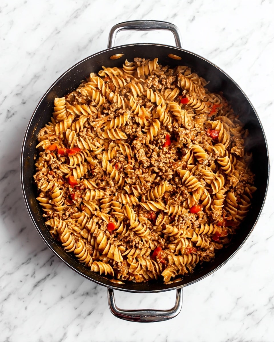A large black pan with two silver handles holds a hot dish of spiral pasta mixed with ground meat and small pieces of red bell pepper. The pasta is light brown, coated with a sauce, and intertwined with the browned meat. Steam rises gently from the pan, showing it is freshly cooked. The pan is set on a white marbled surface. photo taken with an iphone --ar 4:5 --v 7