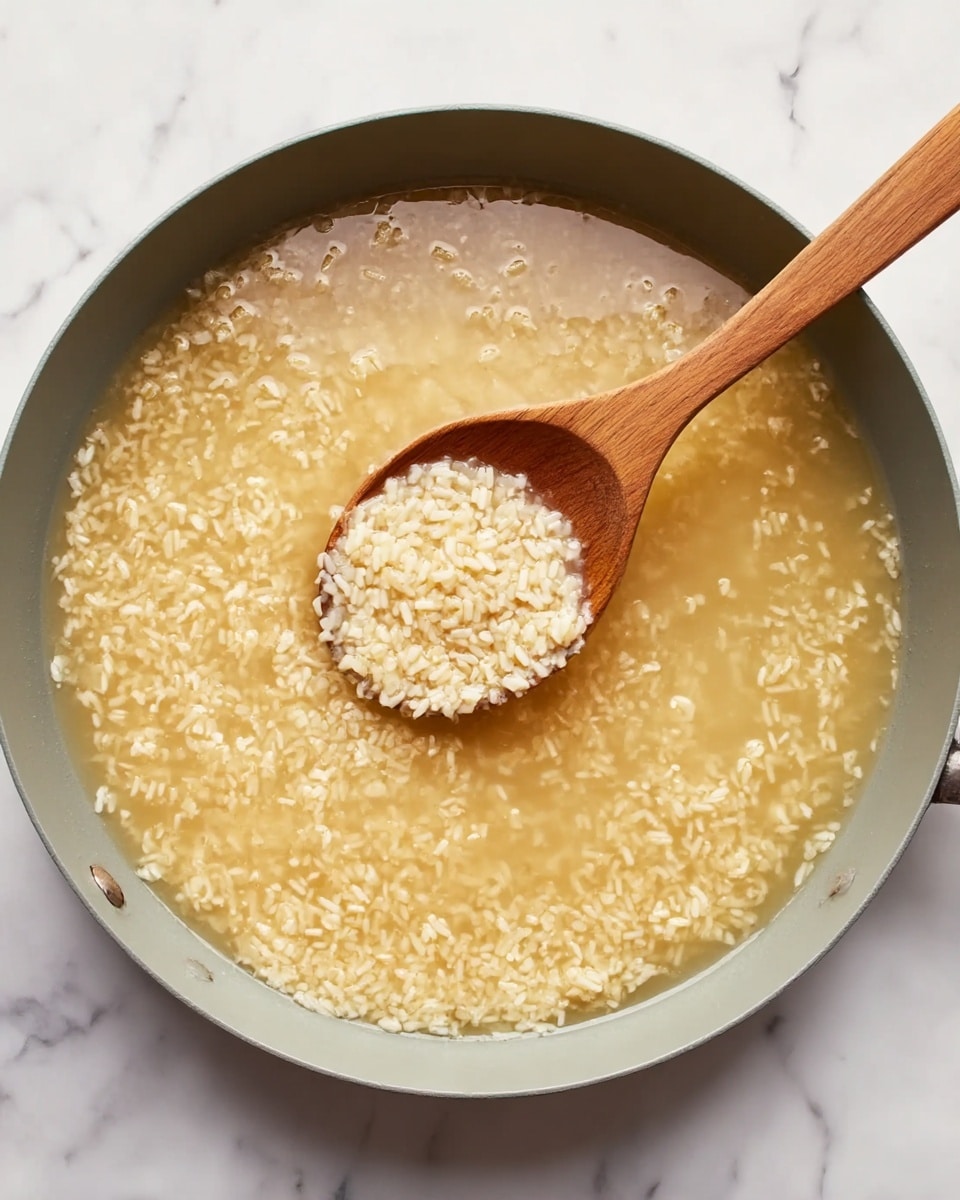 A large round gray pan filled with light golden rice simmering in clear broth, showing many small grains floating and softening. A wooden spoon with a long handle rests inside the pan, lifting some rice, its grains sticky and shining with moisture. The pan sits on a surface with a white marbled texture, adding brightness and contrast to the warm colors inside the pan. The scene looks cozy and simple with natural lighting. photo taken with an iphone --ar 4:5 --v 7