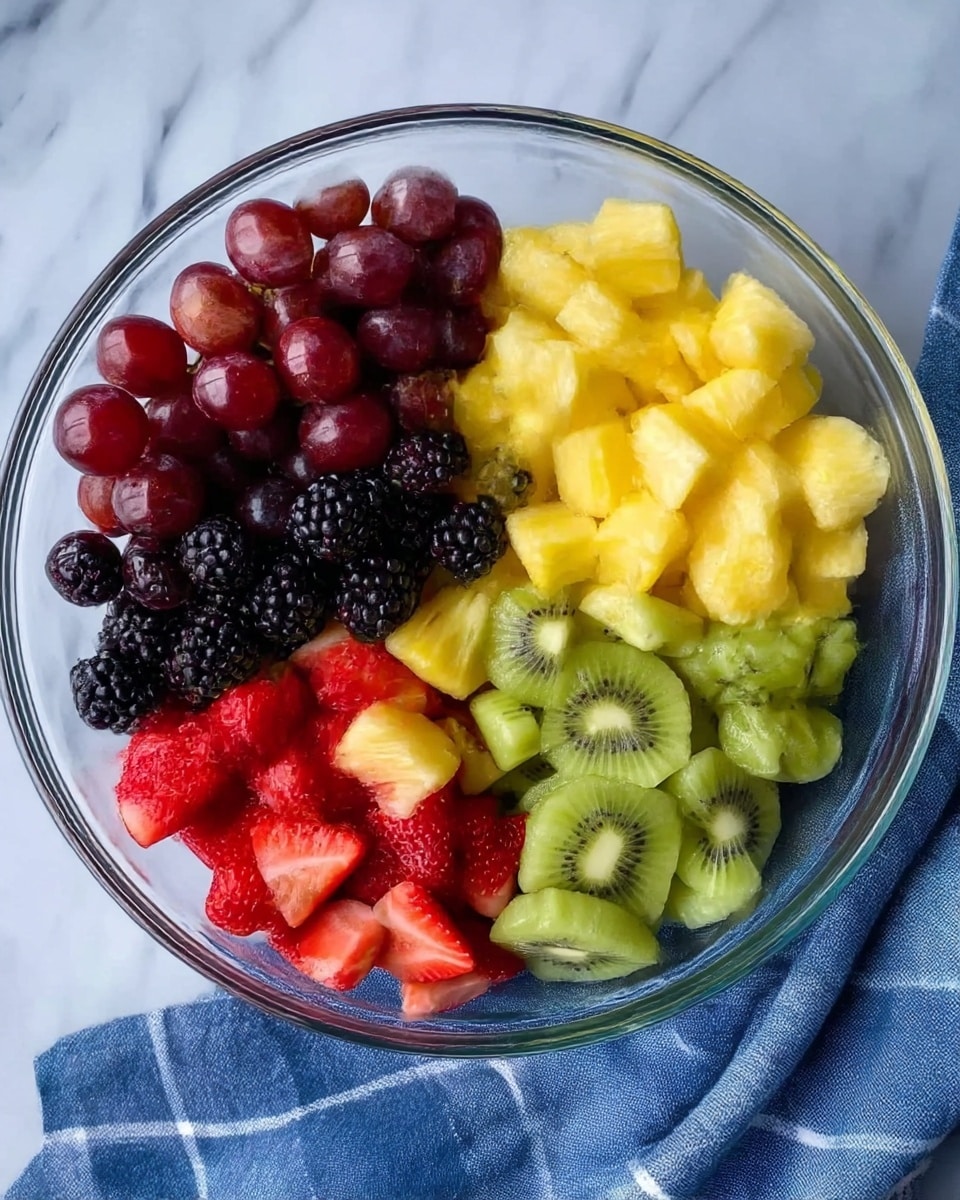 A clear glass bowl filled with five sections of fresh fruit: dark red grapes in one section, blackberries with a bumpy texture next to them, bright yellow pineapple chunks with a juicy look, green slices of kiwi with visible seeds, and red strawberries cut into smaller pieces showing their bright interior. The bowl is placed on a white marbled surface with a blue and white checkered cloth partly visible below. photo taken with an iphone --ar 4:5 --v 7