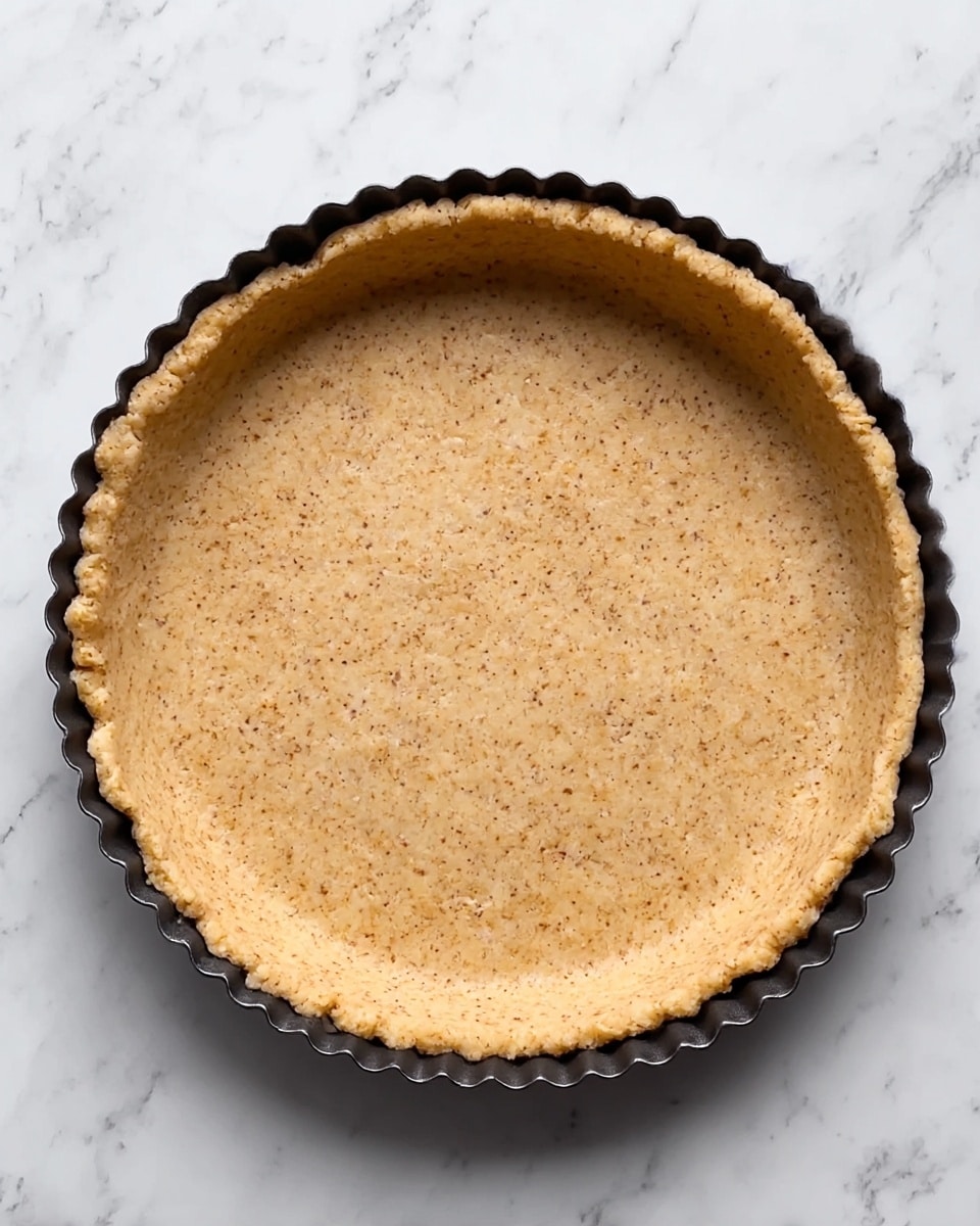 The image shows a single-layer pie crust pressed evenly into a round tart pan with fluted edges. The crust is light brown with small darker specks distributed throughout, giving it a slightly grainy texture. The edges are gently curved upwards to form a shallow wall, and the surface is smooth but not perfectly flat, showing small indentations likely from pressing the dough. The tart pan is dark, with a scalloped rim, sitting on a white marbled surface. photo taken with an iphone --ar 4:5 --v 7