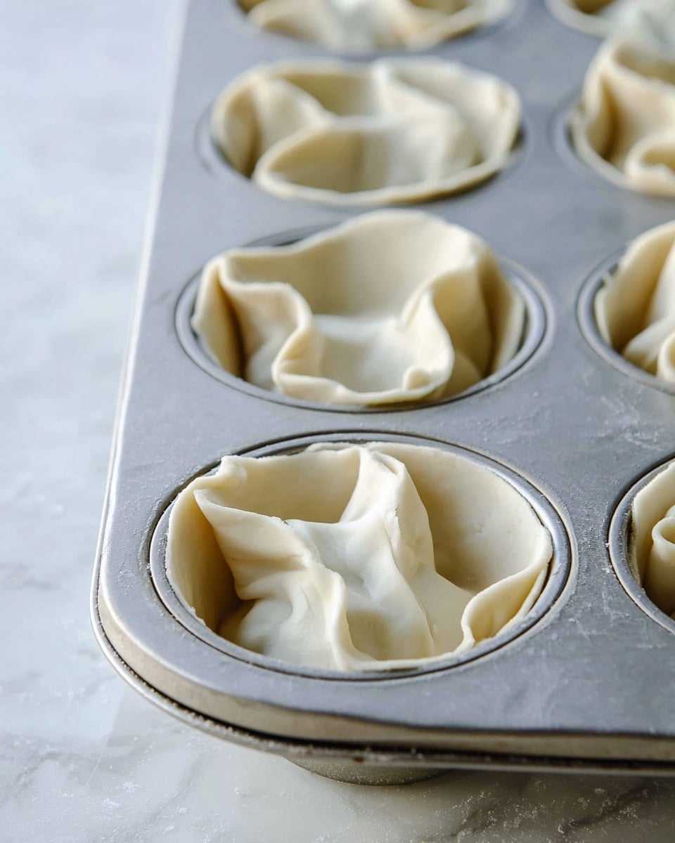 The image shows a close-up of a metal muffin tray with six slots, each lined with a single layer of smooth, pale dough. The dough is placed loosely inside each slot, creating soft folds and gentle waves that rise above the tray's edges. The surface of the dough has a slightly translucent texture with a light, creamy color. The tray sits on a white marbled texture, adding a clean and bright look to the scene. photo taken with an iphone --ar 4:5 --v 7