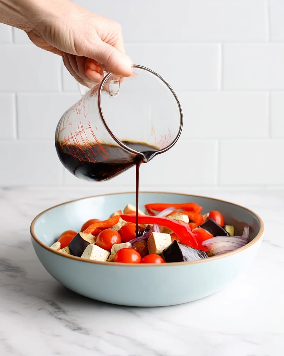 A woman's hand is holding a clear glass measuring cup pouring dark sauce over a bowl of fresh vegetables. The bowl is white and filled with bright red cherry tomatoes, red bell pepper strips, light beige tofu cubes, dark purple eggplant pieces, and sliced red onions, arranged together showing a mix of colors and textures. The bowl sits on a white marbled surface with a clean white tiled wall background. Photo taken with an iphone --ar 4:5 --v 7