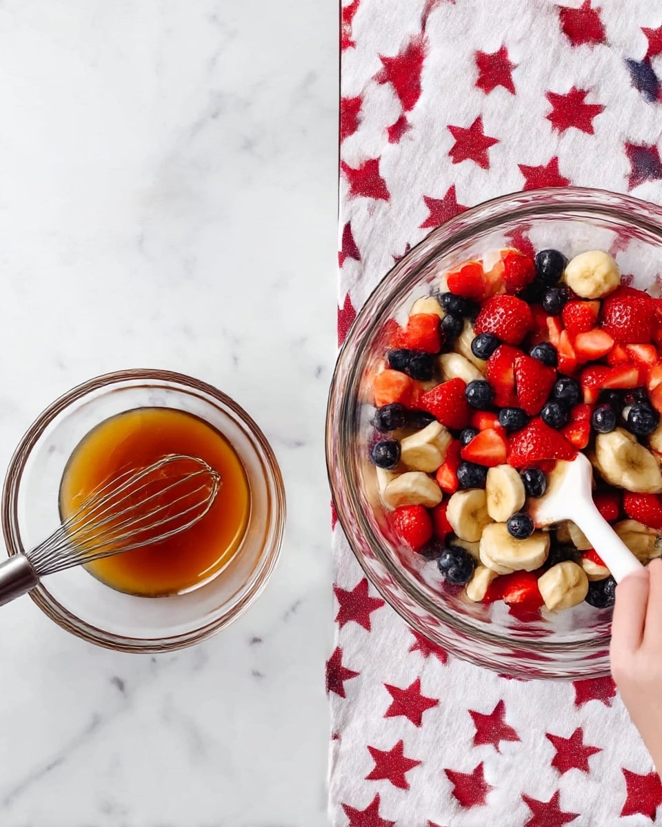 In a clear glass bowl on a white marbled surface with a white and red star-patterned cloth beside it, the scene shows two stages of a fruit salad preparation. On the left, there is a small clear glass bowl filled with a golden-brown liquid, next to a metal whisk with a handle partially visible, resting on the patterned cloth. On the right, a larger clear glass bowl contains three layers of fruit: at the bottom, bright red strawberries cut into halves; mixed throughout are round slices of pale yellow banana and small dark blue blueberries; all mixed together with a white spatula held by a woman's hand gently stirring the fruit. Photo taken with an iphone --ar 4:5 --v 7