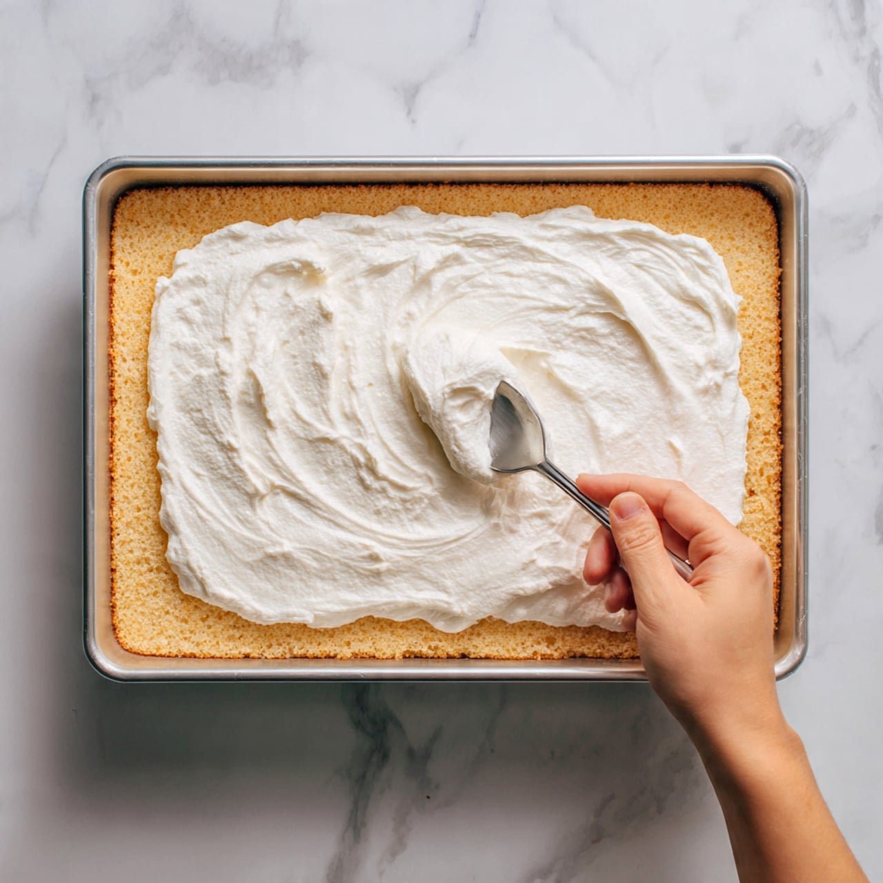 The image shows a baking pan with two distinct layers. The bottom layer is golden brown and textured, filling the whole pan but about one third is uncovered. The top layer is a thick, smooth, white cream spread over most of the pan, with visible soft swirls from spreading. On the right side, a woman's hand holds a spoon covered in the white cream, spreading it over the golden layer. The pan rests on a white marbled surface. Photo taken with an iphone --ar 4:5 --v 7