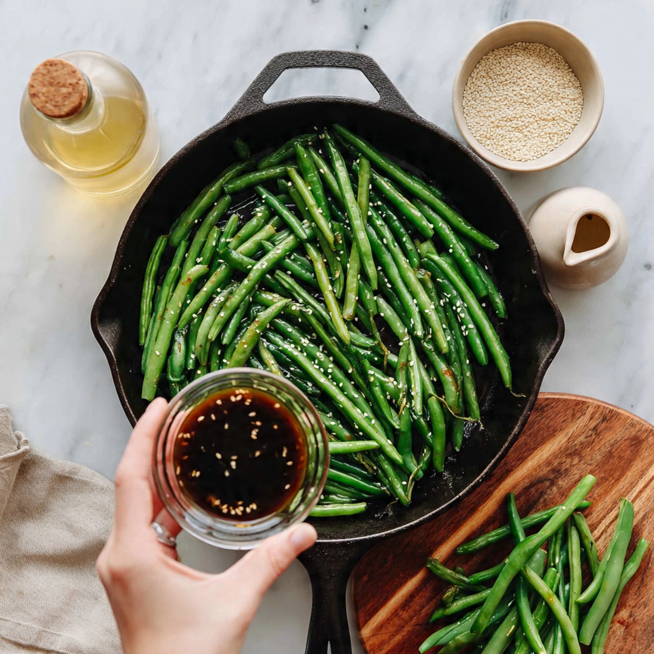 A black cast iron pan filled with bright green cooked green beans sits on a white marbled surface. A woman's hand is holding a small clear glass bowl filled with a dark, thick sauce above the green beans, about to pour it in. Around the pan on the surface are a small clear bottle with light yellow liquid, a beige small plate filled with white sesame seeds, a beige small jug, and a wooden round board with more green beans on it. photo taken with an iphone --ar 4:5 --v 7
