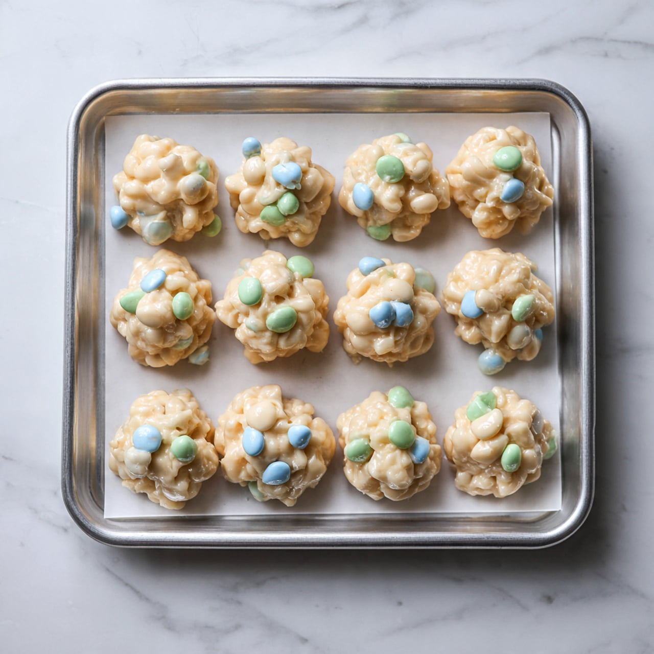 A silver baking tray lined with white parchment paper holds 12 small, round clusters of a light, creamy mixture. Each cluster has a mix of pale beige, light blue, and light green bits spread throughout, giving a bumpy and uneven texture. The clusters are arranged in three rows, with four clusters in each row, spaced evenly but not perfectly round, looking soft and sticky. The scene is set on a white marbled surface. photo taken with an iphone --ar 4:5 --v 7