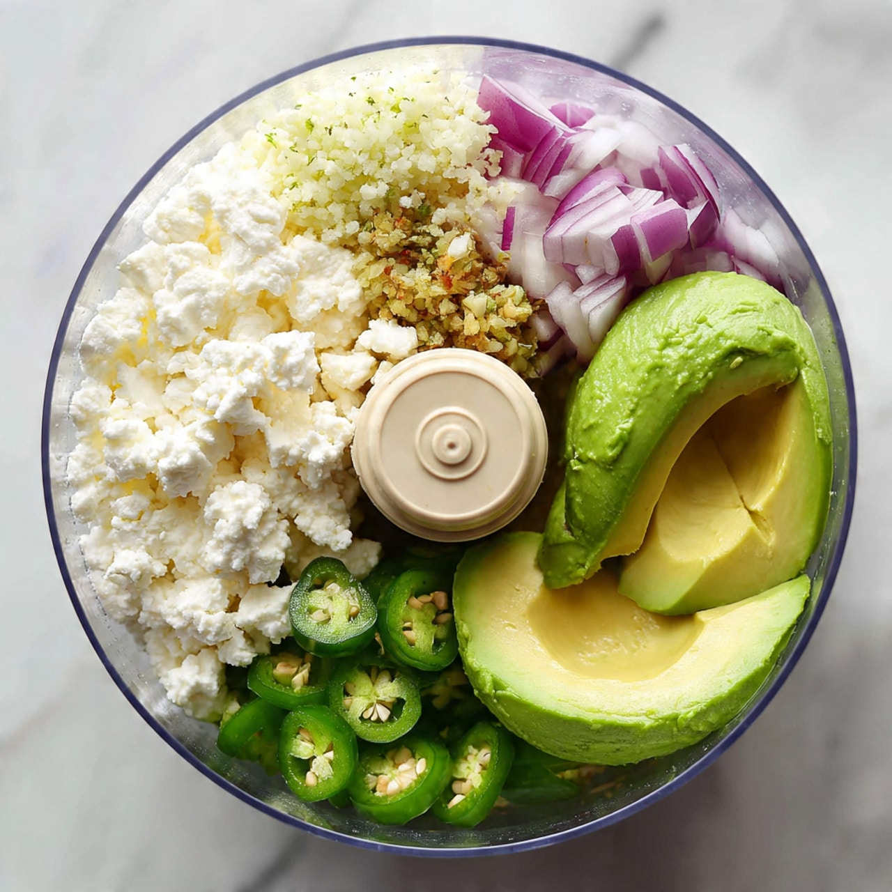 The image shows the inside of a clear food processor bowl filled with several fresh ingredients. On the right side, there are three bright green avocado halves with smooth creamy texture. Above them, chopped green jalapeño peppers with visible seeds and small pieces of purple-red onion are layered closely, adding vibrant color. To the left, there is a generous amount of white cottage cheese with a soft and lumpy texture. Above the cottage cheese, a small heap of minced garlic with a slightly yellow color is placed. All the ingredients are arranged around the central beige food processor blade holder. The background features a white marbled surface. Photo taken with an iphone --ar 4:5 --v 7