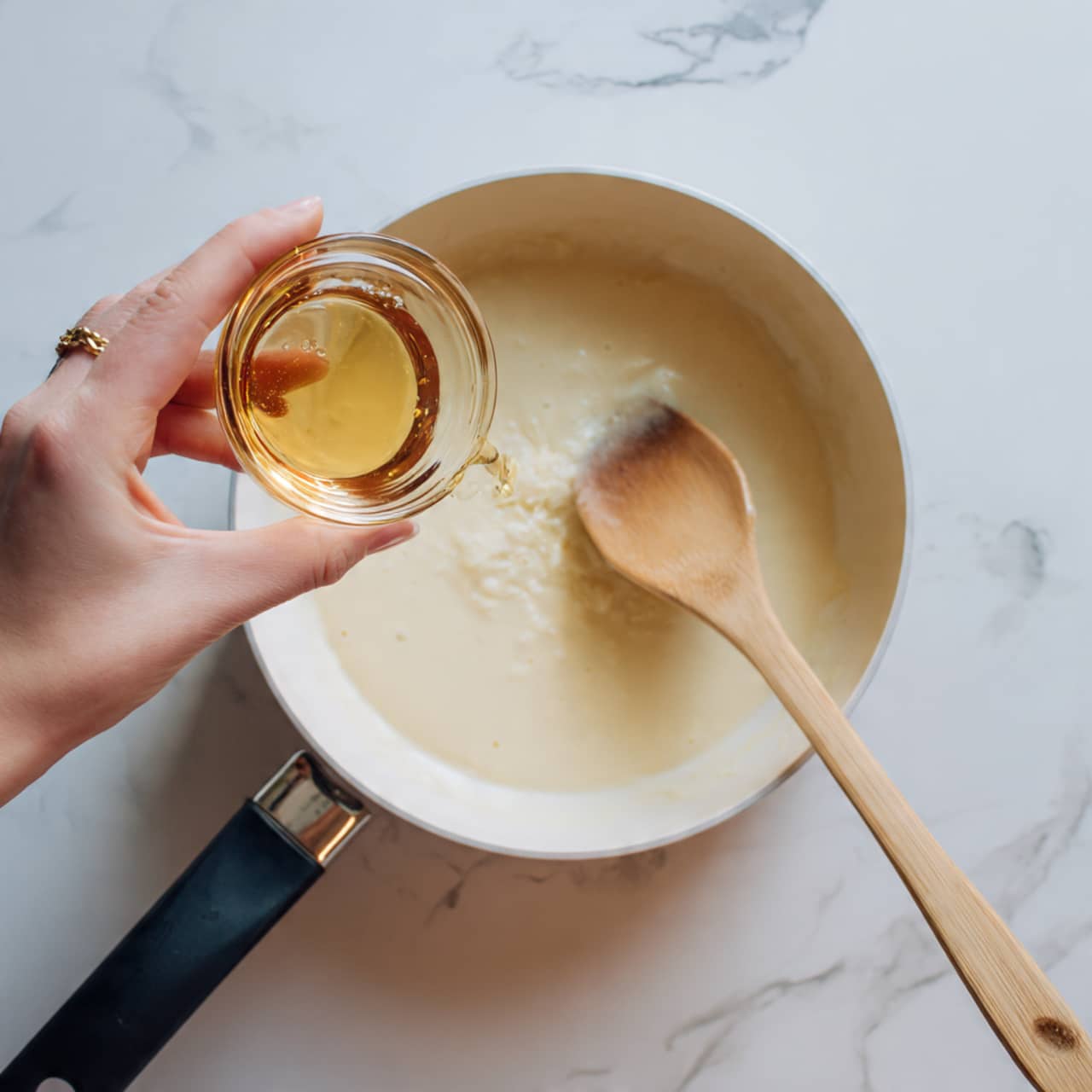 A close-up image shows a woman's hand holding a small glass bowl of light brown liquid, pouring it into a pan filled with a creamy, pale beige mixture. A wooden spoon with a smooth texture is placed inside the pan, stirring the mixture. The pan is set on a white marbled surface, and the scene is lit naturally, highlighting the creamy texture of the mixture and the glossy finish of the wooden spoon. photo taken with an iphone --ar 4:5 --v 7