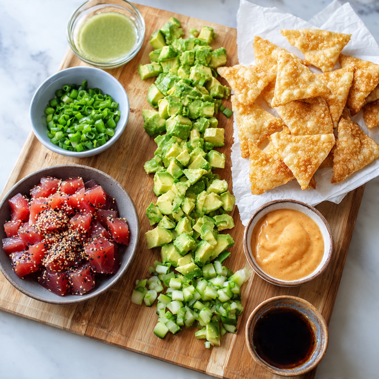 This image shows a wooden board with three piles of chopped ingredients from left to right: green avocado pieces, light green cucumber chunks, and bright green sliced green onions. Below the board, there are five small bowls on a white marbled surface. A gray bowl with dark red raw tuna cubes mixed with spices and black sesame seeds is in the bottom left. To its right, a small brown bowl shows an orange creamy sauce. Above that, a clear bowl holds a light green creamy sauce. Next to it, a small clear bowl has a dark brown liquid, and a tiny brown bowl contains sesame seeds. On the right side, a white plate covered with paper towels holds many golden brown, crispy triangular chips stacked unevenly. Photo taken with an iphone --ar 4:5 --v 7