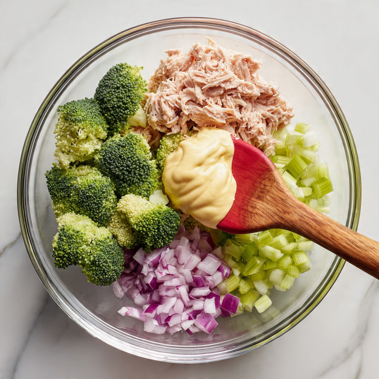 A clear glass bowl sits on a white marbled surface, holding four main ingredients in separate sections: green broccoli florets on the bottom left, light pink tuna flakes on the top middle, light green chopped celery on the top right, and small chopped purple onions on the bottom right. On top of the broccoli, a smooth, creamy pale yellow sauce or mayonnaise is being mixed with a wooden spatula that has a red silicone tip. The ingredients are arranged neatly and not yet mixed. photo taken with an iphone --ar 4:5 --v 7