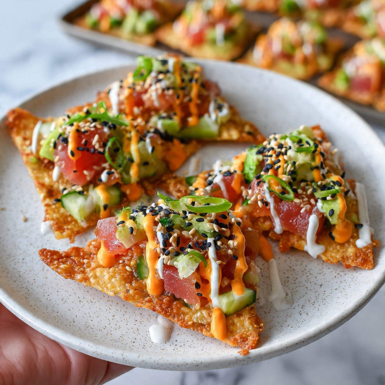 Three crispy triangular chips are arranged on a round white plate with a subtle speckled pattern, sitting on a white marbled surface. Each chip is topped with small chunks of red tuna, bright green cucumber pieces, small bits of avocado, and chopped green onions. The toppings are drizzled with orange and white creamy sauces, and black and white sesame seeds are sprinkled on top, adding texture. The chips have a light golden-brown color with some bubbling on the surface. In the blurred background, a tray with more loaded chips can be seen. Photo taken with an iphone --ar 4:5 --v 7