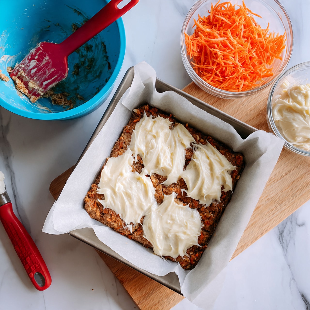 The image shows a square baking pan lined with white parchment paper, filled with one layer of a light brown cake or batter mixed with bits of orange, possibly shredded carrots. On top of this base, there are uneven dollops of creamy white cheese spread over the surface. The pan is placed on a white marbled surface. Next to the pan on the left side, there is a blue mixing bowl with remnants of a brownish batter and a red spatula inside. On the right side, a light wooden cutting board holds two beaters coated lightly with white cream, and behind it is a clear bowl filled with shredded orange carrots. Photo taken with an iphone --ar 4:5 --v 7