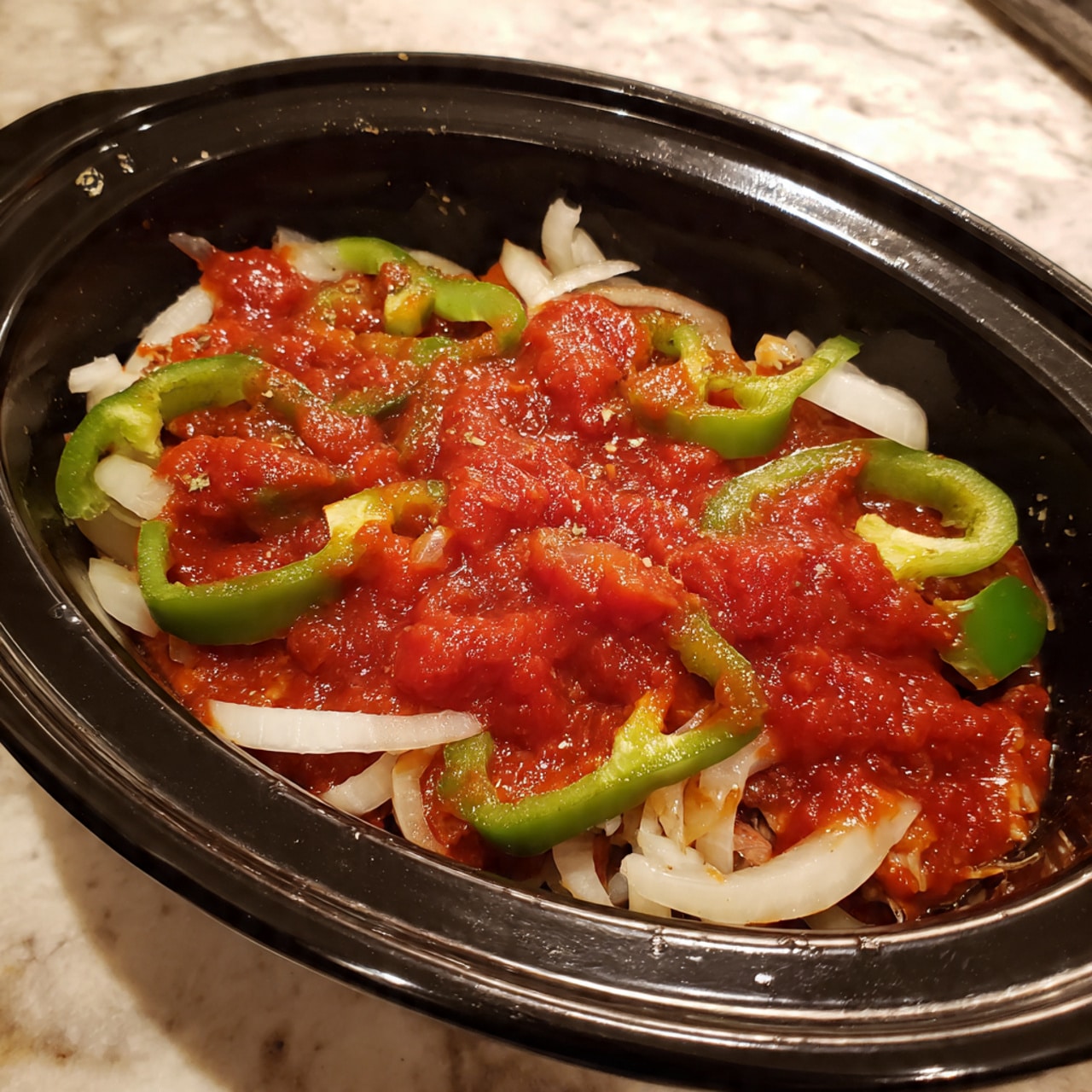 The image shows a black slow cooker filled with raw ingredients layered inside. At the bottom, there are thick white strips of onion spread evenly. On top, there are pieces of raw meat placed across, which are covered generously with bright red tomato sauce. Green pepper slices are scattered throughout the layers, adding a pop of green color. The slow cooker is on a white marbled surface. The photo looks clear and bright, taken with an iphone --ar 4:5 --v 7
