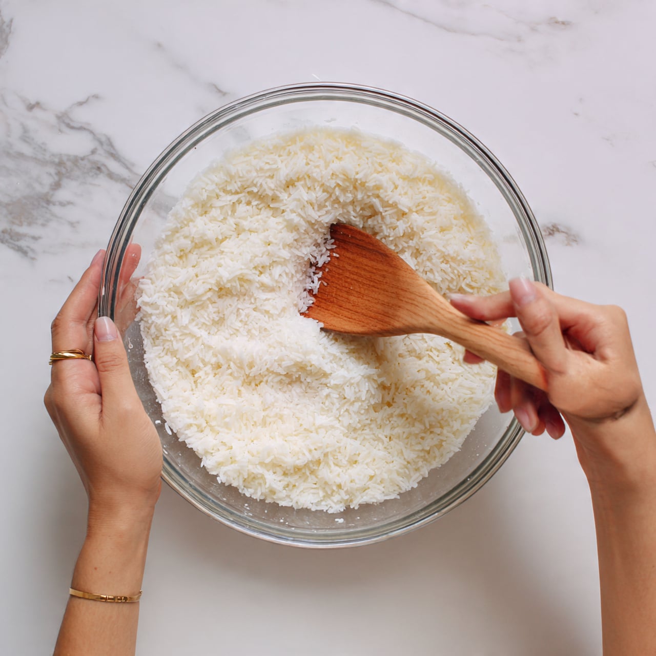 A clear glass bowl filled with a single layer of plump white rice soaked in water. The rice looks soft and slightly separated, with a wooden spatula stirring it gently from the top. Two woman's hands are holding the bowl and the spatula, with light skin and a gold ring visible on one finger. The bowl is placed on a white marbled surface that has faint grey veins, adding a clean and simple background. Photo taken with an iphone --ar 4:5 --v 7