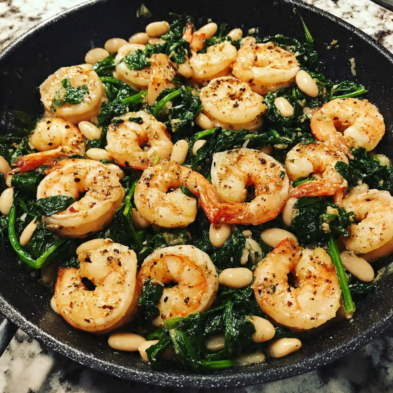 The image shows a close-up of a pan filled with cooked shrimp, wilted dark green spinach leaves, and small white beans spread evenly throughout. The shrimp are light pink with a slightly shiny texture, curled and scattered in a random pattern, mixed well with the leafy spinach and beans. The spinach looks soft and slightly wet, contrasting with the creamy white beans. The pan's dark color makes the shrimp, spinach, and beans stand out clearly. The background is a white marbled texture. photo taken with an iphone --ar 4:5 --v 7