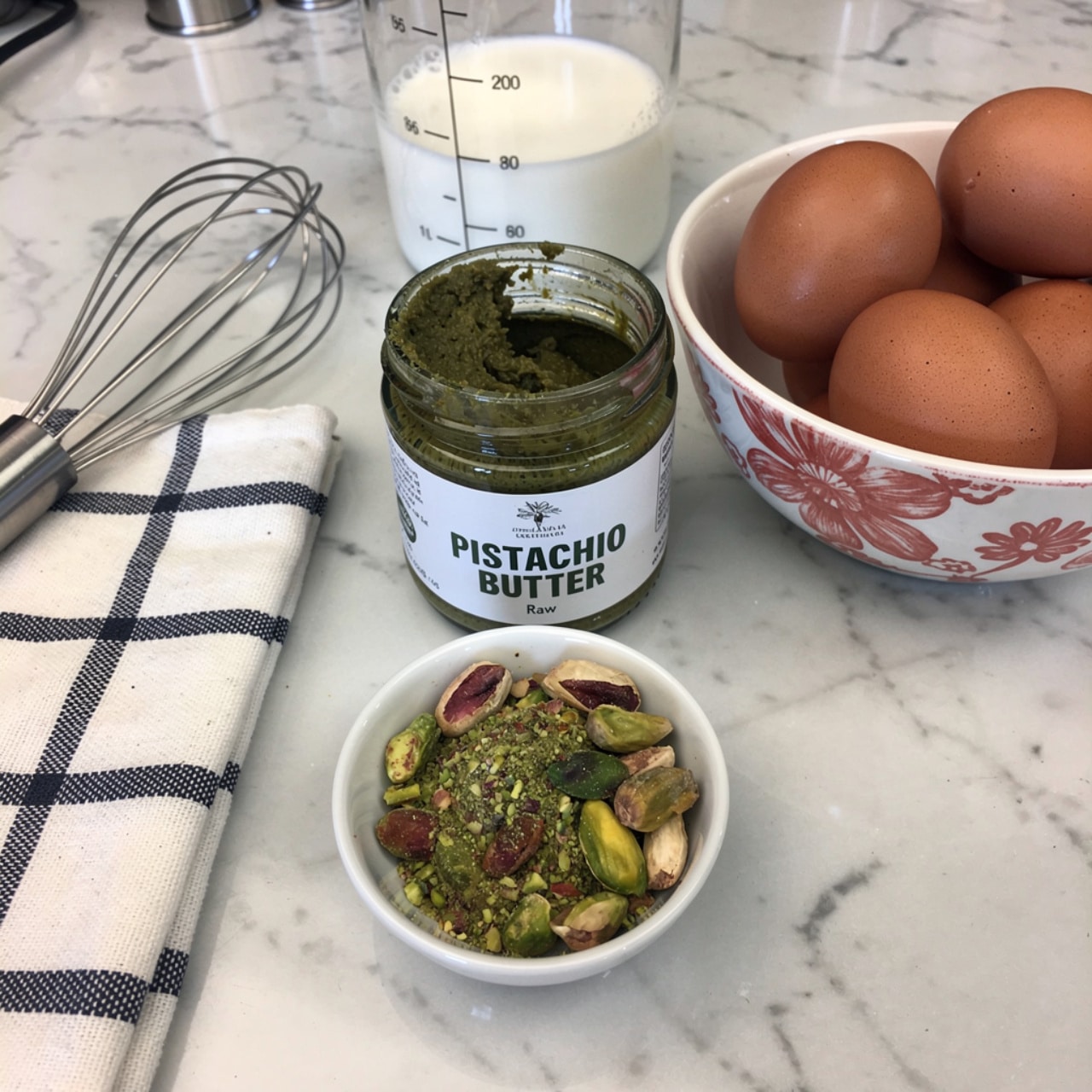 The image shows a white marbled surface with a small open jar of dark green pistachio butter labeled
