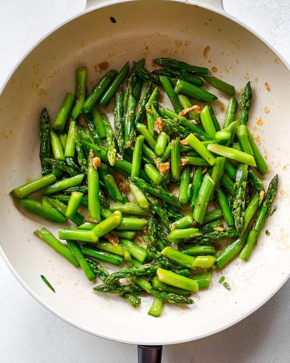 A white pan filled with bright green asparagus pieces that are cut into short, uneven lengths, some showing a slight shine from cooking oil. The asparagus has a tender look with a few parts browned. There are small bits of light golden brown garlic scattered among the asparagus sticks. The pan is on a white marbled surface. photo taken with an iphone --ar 4:5 --v 7