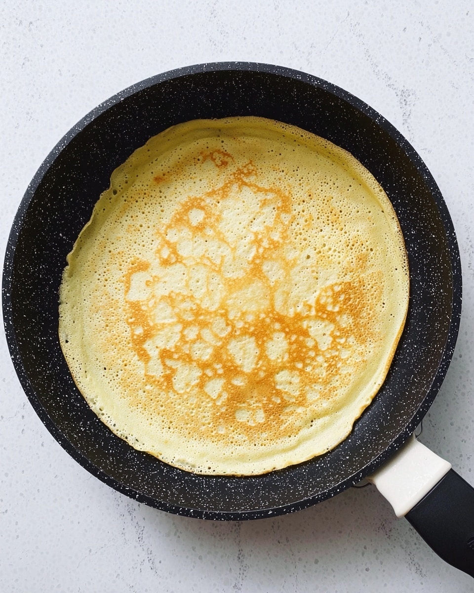 A thin, round crepe with a light golden yellow color and slightly darker golden patches rests flat inside a black speckled frying pan with a white handle. The crepe shows a delicate lacy pattern of small bubbles and soft textures across its surface. The pan is placed on a white marbled surface, giving a clean and minimal background. photo taken with an iphone --ar 4:5 --v 7