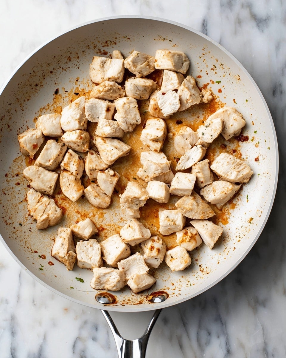 A white pan with a silver handle holds many small pieces of cooked chicken that are light beige with some golden brown edges. The chicken pieces are scattered around the pan with some space between them, showing the browned and slightly oily surface of the pan beneath, which has dark brown marks from cooking. The pan is placed on a white marbled surface. photo taken with an iphone --ar 4:5 --v 7