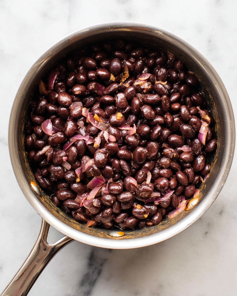 A silver pot filled with cooked black beans mixed with small pieces of cooked red onion, showing a soft and moist texture with the beans glistening slightly. The beans and onions are evenly spread inside the pot with some browning visible on the sides of the pot. The pot is placed on a white marbled surface, and the photo is taken from above, capturing the round shape of the pot and its handle extending upwards. Photo taken with an iphone --ar 4:5 --v 7