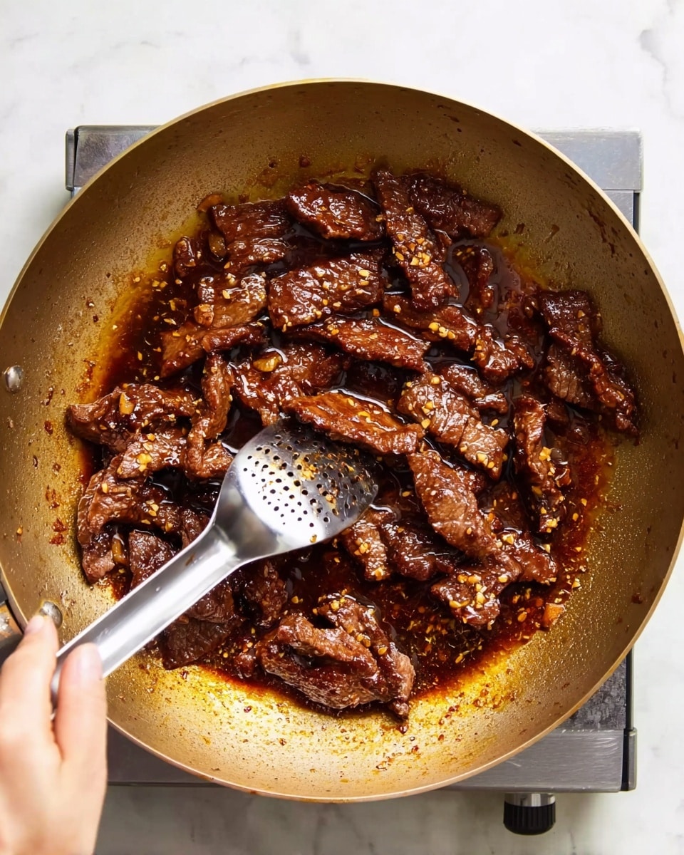 A close-up view of a large metal wok filled with cooked dark brown beef strips in a glossy, oily sauce with small bits of garlic or spices visible. The beef pieces have a rich, caramelized texture and irregular shapes. A woman's hand is holding a shiny metal slotted spoon, stirring the beef gently. The wok sits on a small portable stove on a white marbled surface, and the light creates slight reflections on the meat and oil, emphasizing the sizzling effect. photo taken with an iphone --ar 4:5 --v 7